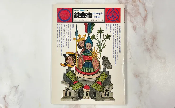 錬金術の象徴的なイラストが描かれた『錬金術のイメージの博物誌』の表紙。王冠をかぶった人物とその隣に立つ人物、植物、フクロウ、建物が描かれている。上部には日本語のタイトルと説明がある。